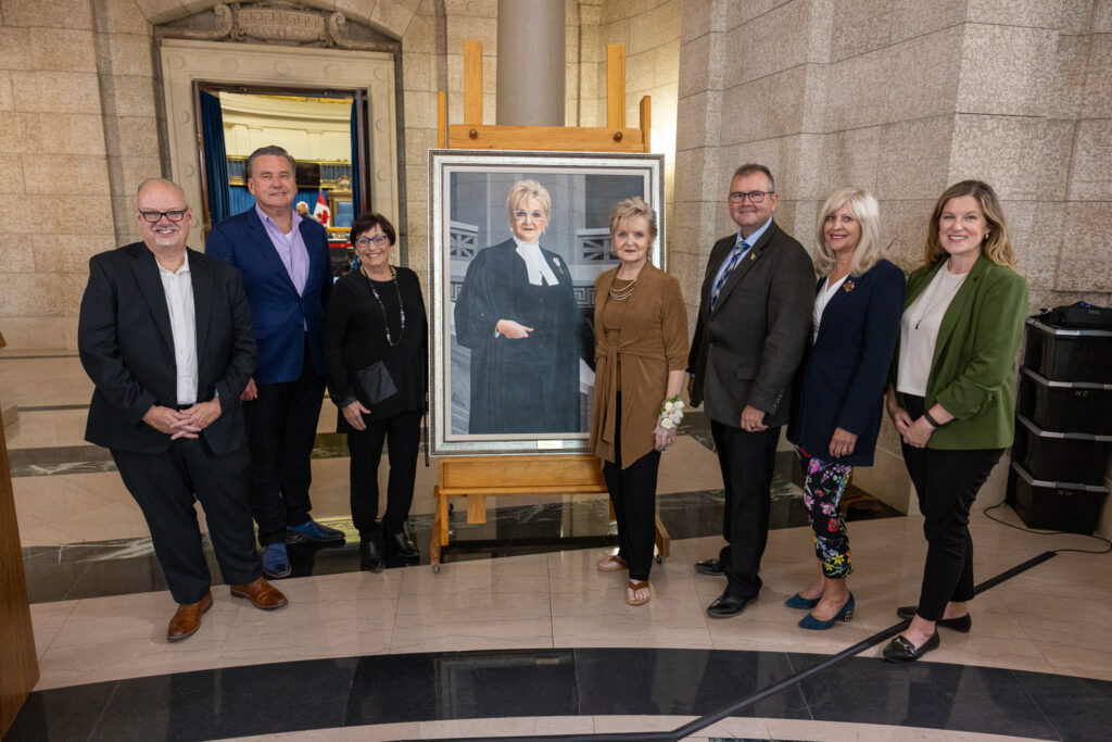 Myrna Driedger and her portrait, with MLA Kelvin Goertzen, MLA Doyle Piwniuk, former PC MLA Bonnie Mitchelson, former Speaker Myrna Driedger, Opposition Leader Wayne Ewasko, former PC MLA Cathy Cox, and MLA Kathleen Cook